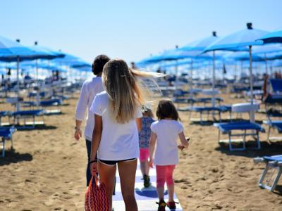 Famiglia cammina sulla spiaggia tra ombrelloni blu e sedie a sdraio.