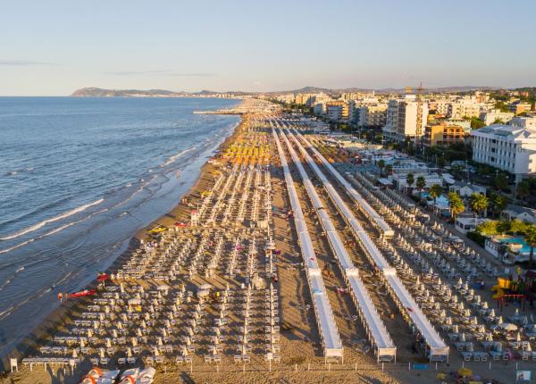 Spiaggia affollata con ombrelloni e mare calmo, città sullo sfondo.